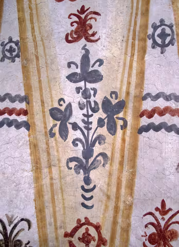 Pictură bisericească de perete: pilaștri ocru și un buchet floral central albastru-gri, cu benzi ondulate și rozete.